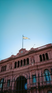 An ornate pink building with classical architectural elements, featuring multiple arches, columns, and decorative sculptures. A clock is centered at the top, and an Argentine flag waves proudly above. The sky is clear and bright.