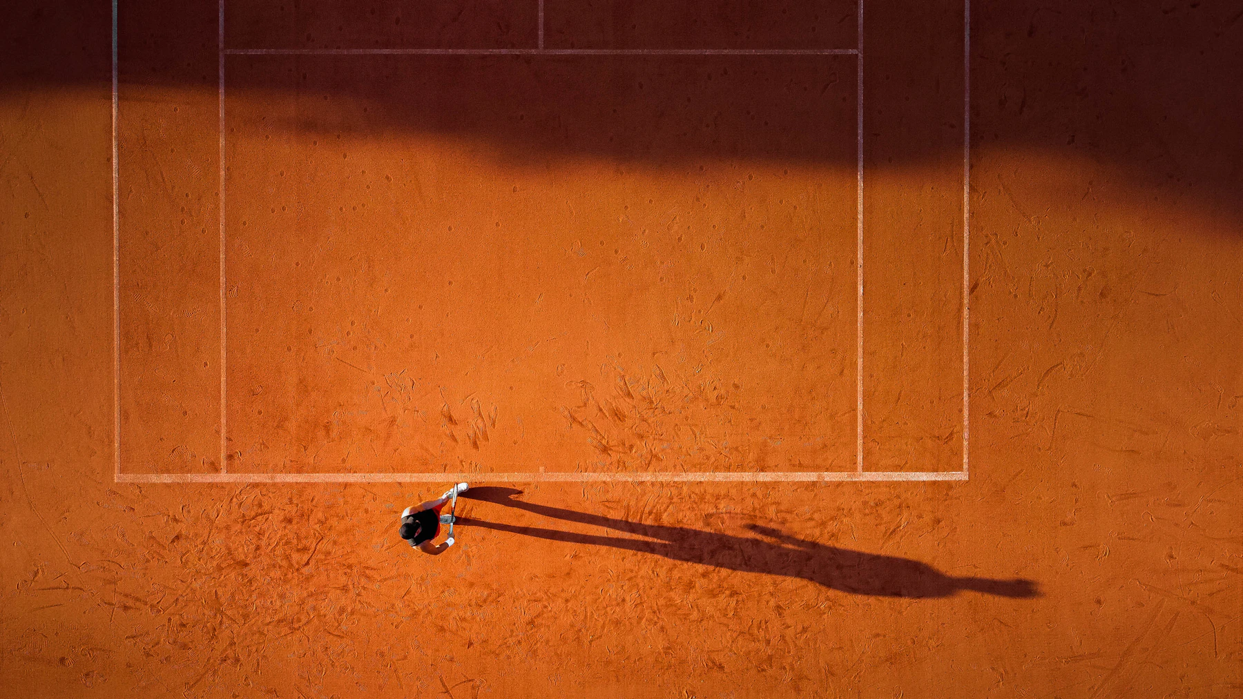 A red clay tennis court from above, white lines, a single player's long shadow