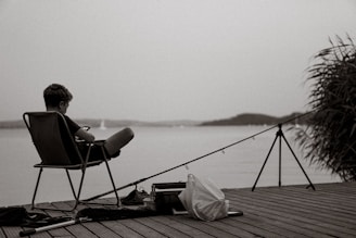 A person sitting on a folding chair on a wooden dock, with their feet up and holding a smartphone. A fishing rod is set up beside them, and a plastic bag and tackle box are on the dock. The background features a calm, expansive body of water and distant hills, with some grasses visible on the right side.