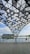A geometric steel canopy with perforated triangular patterns forms a shaded structure over a wooden deck. Beyond the railing, a calm body of water reflects the blue sky and scattered clouds. In the distance, there is a modern architectural building and lush greenery along the horizon.