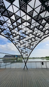 A geometric steel canopy with perforated triangular patterns forms a shaded structure over a wooden deck. Beyond the railing, a calm body of water reflects the blue sky and scattered clouds. In the distance, there is a modern architectural building and lush greenery along the horizon.