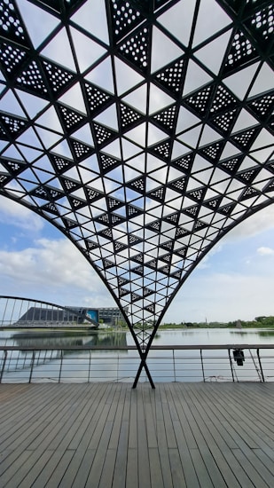 A geometric steel canopy with perforated triangular patterns forms a shaded structure over a wooden deck. Beyond the railing, a calm body of water reflects the blue sky and scattered clouds. In the distance, there is a modern architectural building and lush greenery along the horizon.