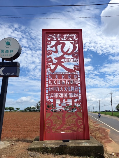 A large red decorative sign with Chinese characters and the word 'TAIWAN' prominently displayed. The sign is set against a backdrop of a blue sky with scattered clouds. To the left, there is a signpost with some writing in a turquoise and white circular sign. A road runs alongside a brown plowed field on the right, where a person on a motorcycle is visible.