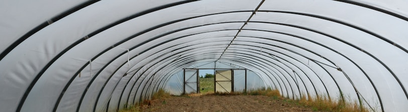 A long, empty tunnel-shaped greenhouse with a curved metal frame and translucent covering. The inside ground is bare soil with some patches of grass along the sides. In the distance, an open door reveals an outdoor field and sky.
