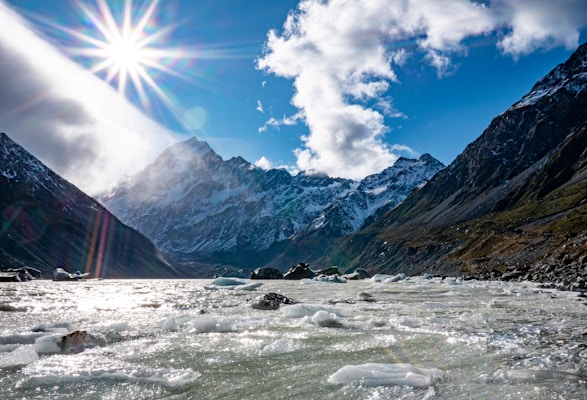 A breathtaking mountain landscape with rugged peaks covered in snow, surrounded by clear blue skies and a bright sun. A glacial river flows in the foreground, with chunks of ice visible on the surface. The sunlight creates a burst of rays and a lens flare effect, adding a dramatic element to the scene.