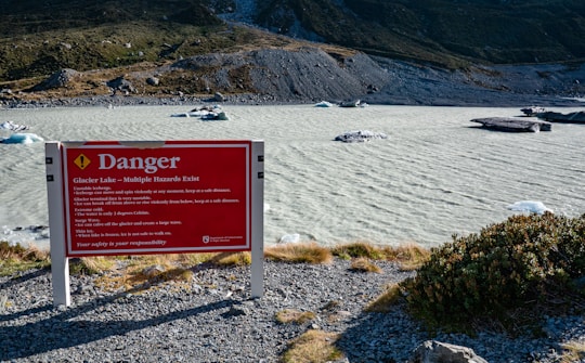 A warning sign is prominently displayed by a glacial lake surrounded by rocky terrain and distant hills. The sign reads 'Danger' and provides warnings about the hazards of unstable icebergs and cold water, highlighting the extreme caution needed in the area. The lake has small icebergs floating on the surface, and the surrounding landscape is rugged and sparsely vegetated.