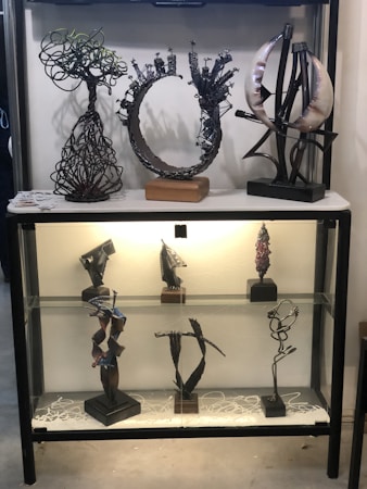 An art display cabinet contains multiple abstract metal sculptures. The upper shelf features three intricate pieces: a tangled tree-like structure, a circular form with small protrusions, and a composition involving curved elements. The lower shelf holds five smaller sculptures, each with unique geometric and organic designs, showcasing abstract artistry.