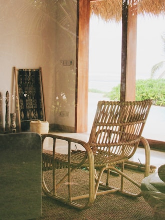 A cozy interior space with a vintage rattan chair positioned on a woven rug. Natural light filters in through a large glass door, leading to an outdoor view with green foliage and a glimpse of the ocean. Decorative wooden items and a rolled-up mat lean against the wall.