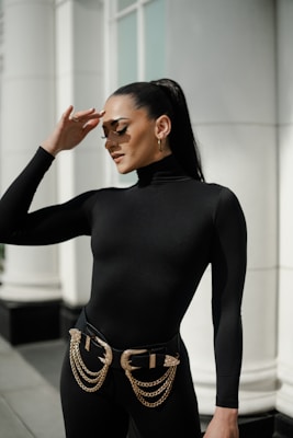 A person wearing a black form-fitting outfit with gold chain belt accessories stands in an outdoor urban setting. The individual has long dark hair tied back in a ponytail and is wearing large fashion sunglasses. The background includes a building with large windows and pillars.