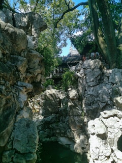 Rocky landscape with large stone formations and lush greenery, featuring a traditional Chinese architectural structure in the background. The area is shaded by tall trees, creating a serene and tranquil atmosphere.