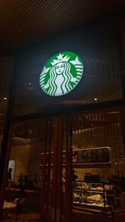 A brightly lit storefront with a circular green and white logo displayed prominently above the entrance. Inside, there is a menu board with various offerings and a counter with pastries on display. The interior features warm lighting and wooden decor.