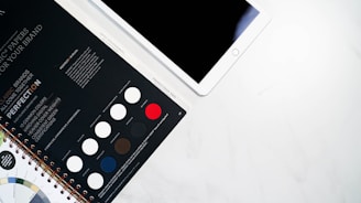 A spiral-bound swatch book featuring various paper options is partially opened next to a tablet with a blank screen lying on a white surface. The swatch book displays different color samples, including white, black, red, and shades of blue and brown, with text detailing paper features and branding options.
