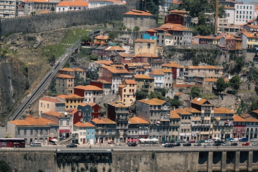 A densely packed hillside cityscape with numerous colorful buildings, each topped with orange tiled roofs. A stone wall runs along the upper part of the hill, and a sloping funicular railway can be seen on the left side. The buildings are set against a backdrop of lush greenery and rugged cliffs. Cars are parked along the road at the bottom, with a few people visible on the sidewalks.