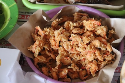 A bowl filled with crispy, fried shrimp, presented on a piece of brown paper which lies on a purple tray. The shrimp have a golden, crunchy batter and are placed in a casual, informal setting.