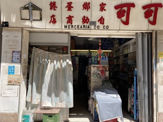 A small convenience store entrance with a curtain partially covering the doorway. The store has a variety of goods including bottled water, drinks, and household items. The signage above the entrance includes both Chinese characters and English text.