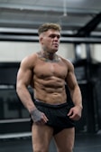 A muscular man with tattoos is standing, showcasing a toned physique. He has short, curly hair and is wearing black athletic shorts. The setting appears to be an indoor gym with exercise equipment in the background.