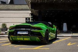 A vibrant green sports car is parked in a lot outside a modern building with wooden siding. The car is sleek with a convertible top and stylish rear design. The license plate is visible, and the setting appears to be well-maintained with neat landscaping.