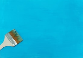 A paintbrush with a wooden handle rests on a bright blue canvas. The bristles appear to have blue paint on them, suggesting recent use.