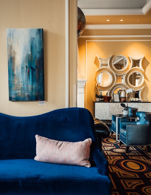 A modern living room features a plush blue velvet sofa adorned with a single soft pillow. The wall behind displays an abstract painting with hints of blue. Adjacent is a stylish area with geometric patterned mirrors and a decorative console table. The lighting is warm and inviting, enhancing the yellowish wall tones.
