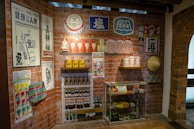 A retro-themed room displays vintage posters and advertisements on brick walls featuring products like drinks and household items. There are shelves with classic bottles, tins, and jars. A glass cabinet contains various collectibles and packaged goods. A traditional woven hat is mounted on the wall.