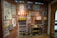 A retro-themed room displays vintage posters and advertisements on brick walls featuring products like drinks and household items. There are shelves with classic bottles, tins, and jars. A glass cabinet contains various collectibles and packaged goods. A traditional woven hat is mounted on the wall.