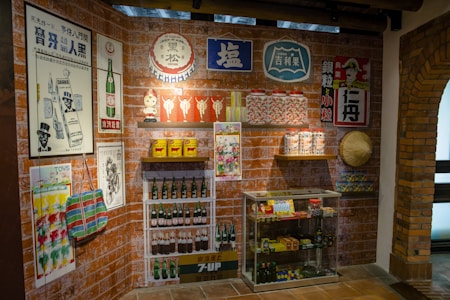 A retro-themed room displays vintage posters and advertisements on brick walls featuring products like drinks and household items. There are shelves with classic bottles, tins, and jars. A glass cabinet contains various collectibles and packaged goods. A traditional woven hat is mounted on the wall.