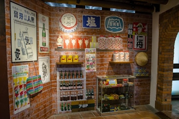A retro-themed room displays vintage posters and advertisements on brick walls featuring products like drinks and household items. There are shelves with classic bottles, tins, and jars. A glass cabinet contains various collectibles and packaged goods. A traditional woven hat is mounted on the wall.