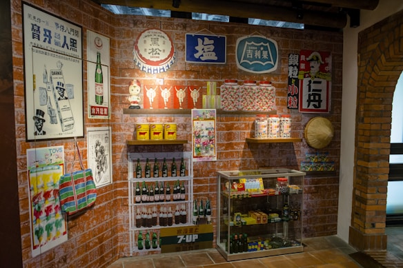 A retro-themed room displays vintage posters and advertisements on brick walls featuring products like drinks and household items. There are shelves with classic bottles, tins, and jars. A glass cabinet contains various collectibles and packaged goods. A traditional woven hat is mounted on the wall.