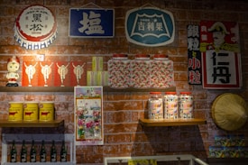 A nostalgic collection display featuring vintage-style items against a brick wall. The display includes several retro advertisements and signs in Chinese and Japanese characters, jars filled with candy, old-fashioned milk tins, a mannequin in a toy box, and a decorative straw hat.