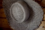 A textured, woven straw hat with a wide brim, resting against a wooden surface. The hat's intricate pattern is highlighted by shadows and light.