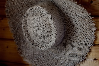 A textured, woven straw hat with a wide brim, resting against a wooden surface. The hat's intricate pattern is highlighted by shadows and light.