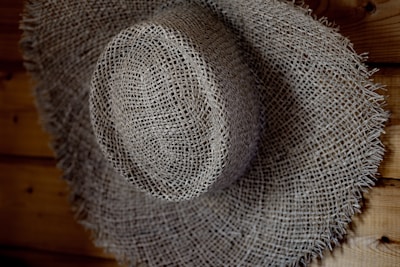A textured, woven straw hat with a wide brim, resting against a wooden surface. The hat's intricate pattern is highlighted by shadows and light.