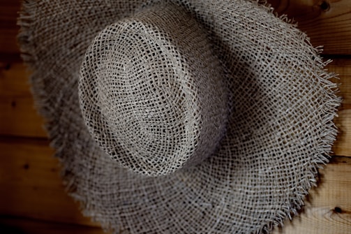 A textured, woven straw hat with a wide brim, resting against a wooden surface. The hat's intricate pattern is highlighted by shadows and light.