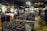 A store filled with heavy metal merchandise, including band shirts, hats, patches, and posters. The walls are adorned with graphic artwork and a variety of metal band logos. A cardboard cutout of a musician stands near a changing area. The store is lit with a chandelier and accented with cymbals on the wall, contributing to the metal-themed decor.