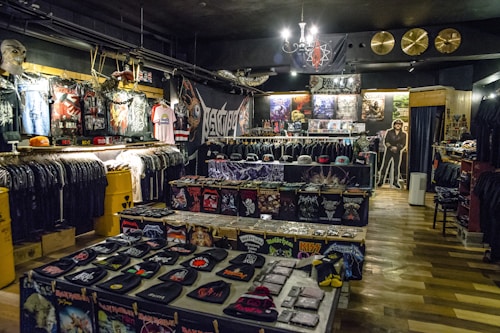 A store filled with heavy metal merchandise, including band shirts, hats, patches, and posters. The walls are adorned with graphic artwork and a variety of metal band logos. A cardboard cutout of a musician stands near a changing area. The store is lit with a chandelier and accented with cymbals on the wall, contributing to the metal-themed decor.