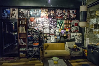 The room is decorated with a variety of band posters covering the wall, featuring heavy metal and rock bands. There's a punching bag hanging from the ceiling, a shelf filled with vinyl records and band merchandise, and a small stage area. A sofa and chairs are placed in front of the posters, and a table is in the center with a small speaker and a skull decoration on it. The atmosphere is cluttered yet cozy, with dim lighting enhancing a rock-and-roll vibe.