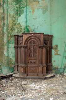 An intricately carved wooden confessional box placed against a weathered, peeling mint-green wall. The floor is covered with debris and rubble, suggesting a state of disrepair or abandonment.