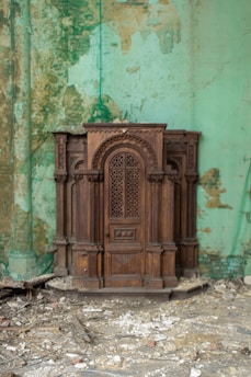 An intricately carved wooden confessional box placed against a weathered, peeling mint-green wall. The floor is covered with debris and rubble, suggesting a state of disrepair or abandonment.