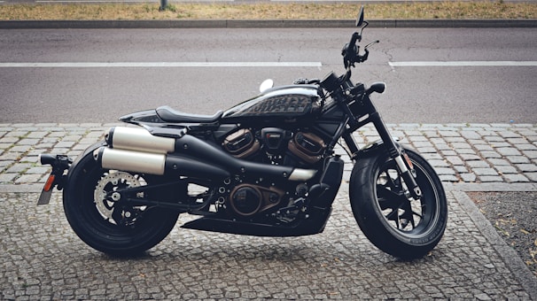 A sleek black motorcycle with metallic accents is parked on a cobblestone pavement beside a street. The motorcycle features prominent exhaust pipes and a modern design, suggesting its suitability for urban environments. The background includes a glimpse of the road and a patch of grass, adding an urban context to the setting.