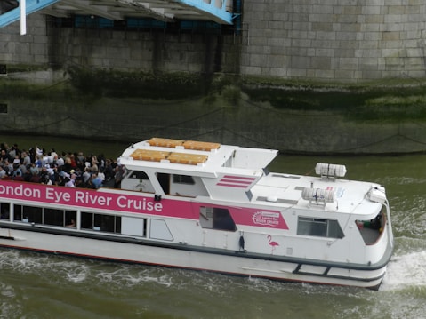 A river cruise boat with a pink and white color scheme navigates a waterway. Numerous passengers are visible on the deck, enjoying the view. The side of the boat has signage for the 'London Eye River Cruise.' The boat is passing under a structure, with a stone wall in the background.