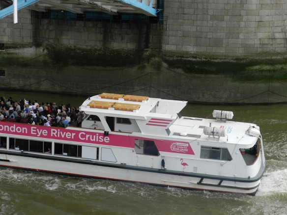 A river cruise boat with a pink and white color scheme navigates a waterway. Numerous passengers are visible on the deck, enjoying the view. The side of the boat has signage for the 'London Eye River Cruise.' The boat is passing under a structure, with a stone wall in the background.
