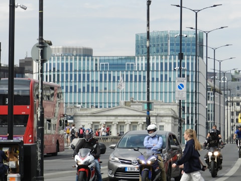 Urban street scene featuring a modern glass building with vertical stripes in the background. A red double-decker bus is moving on the left, and a few motorcyclists with helmets ride through the scene. Several pedestrians, including a person walking on the sidewalk, are visible. Traffic lights, street lamps, and parking signs are present, adding to the busy city atmosphere.