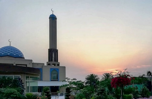 A mosque with a tall minaret and a blue-domed roof is surrounded by lush greenery and flowering plants. The sky is painted with soft hues of orange and yellow, indicating either a sunrise or sunset.