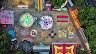 An aerial view of a colorful amusement park, showcasing various rides like a carousel, Ferris wheel, bumper cars, and other attractions spread out over a sandy surface. Brightly colored tents and booths line the perimeter, and lush green trees border the park.