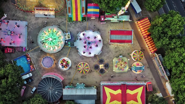 An aerial view of a colorful amusement park, showcasing various rides like a carousel, Ferris wheel, bumper cars, and other attractions spread out over a sandy surface. Brightly colored tents and booths line the perimeter, and lush green trees border the park.