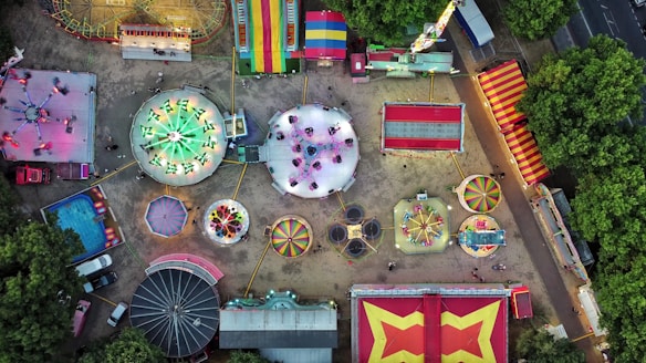 An aerial view of a colorful amusement park, showcasing various rides like a carousel, Ferris wheel, bumper cars, and other attractions spread out over a sandy surface. Brightly colored tents and booths line the perimeter, and lush green trees border the park.