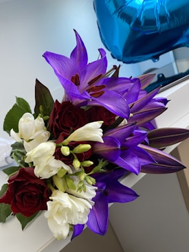 A vibrant bouquet of flowers featuring deep purple lilies, rich red roses, and creamy white blooms, accompanied by lush green leaves. In the background, a shiny blue foil balloon is partially visible, resting on a light-colored surface.