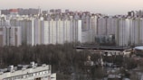 High-rise residential buildings dominate the urban skyline, with rows of identical apartment blocks. The foreground features a park with bare trees, suggesting a winter or early spring setting. A smaller traditional-style structure with a domed roof is nestled among the trees.
