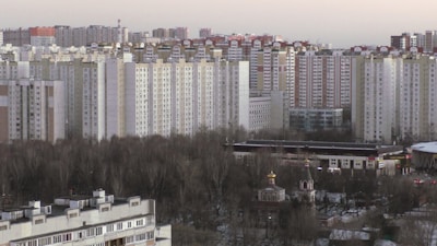 High-rise residential buildings dominate the urban skyline, with rows of identical apartment blocks. The foreground features a park with bare trees, suggesting a winter or early spring setting. A smaller traditional-style structure with a domed roof is nestled among the trees.