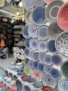 A market stall displays a wide array of brightly colored, intricately patterned ceramic plates, bowls, and pottery. The designs are primarily geometric and floral, featuring dominant hues of blue, red, green, and white. To the left, a person is seated, wearing a hoodie and a cap, with bags and other pottery items in the background.